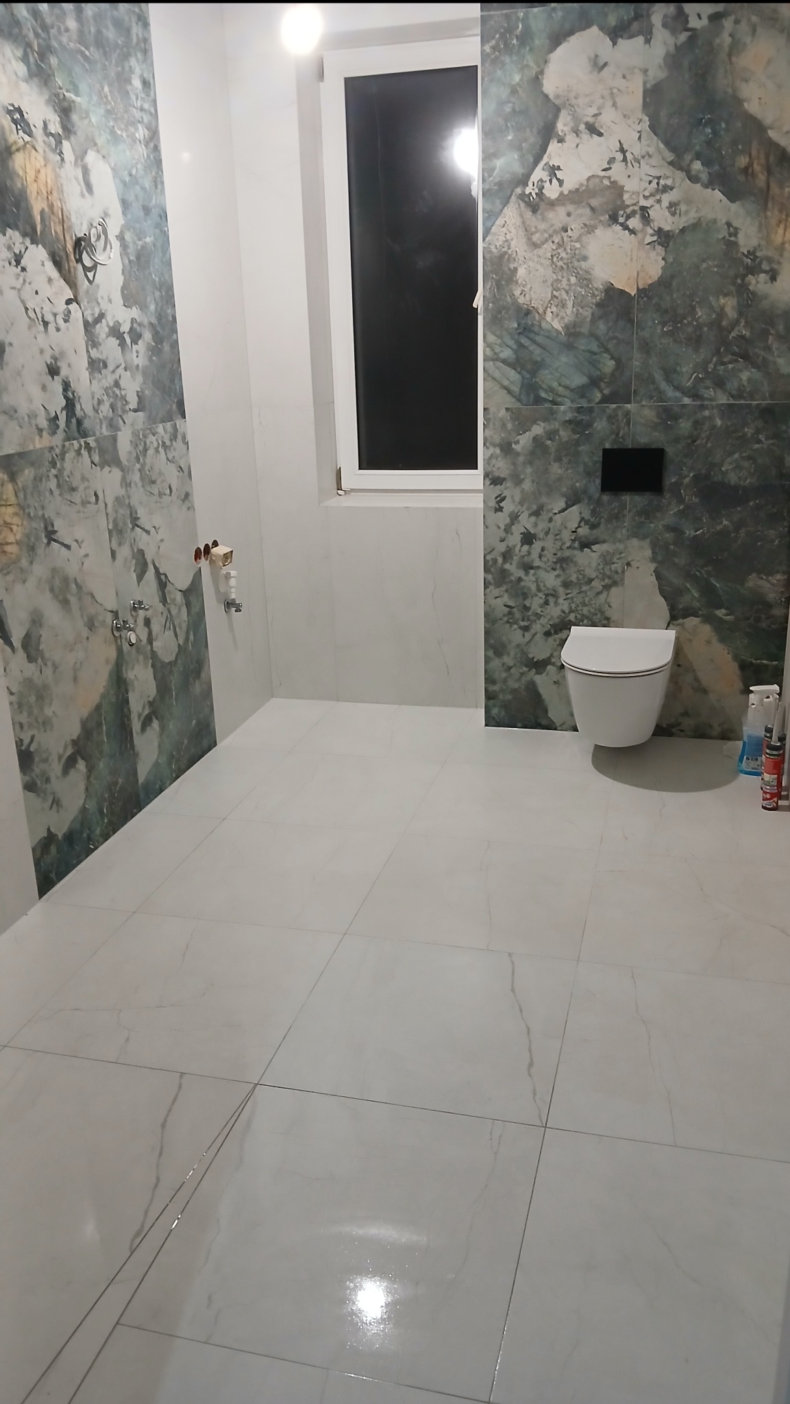 Wykończona łazienka z marmurowymi płytkami na ścianach i podłodze, wisząca toaleta, okno z białymi ramami, armatura. Nowoczesny design z akcentem na naturalne wzory.