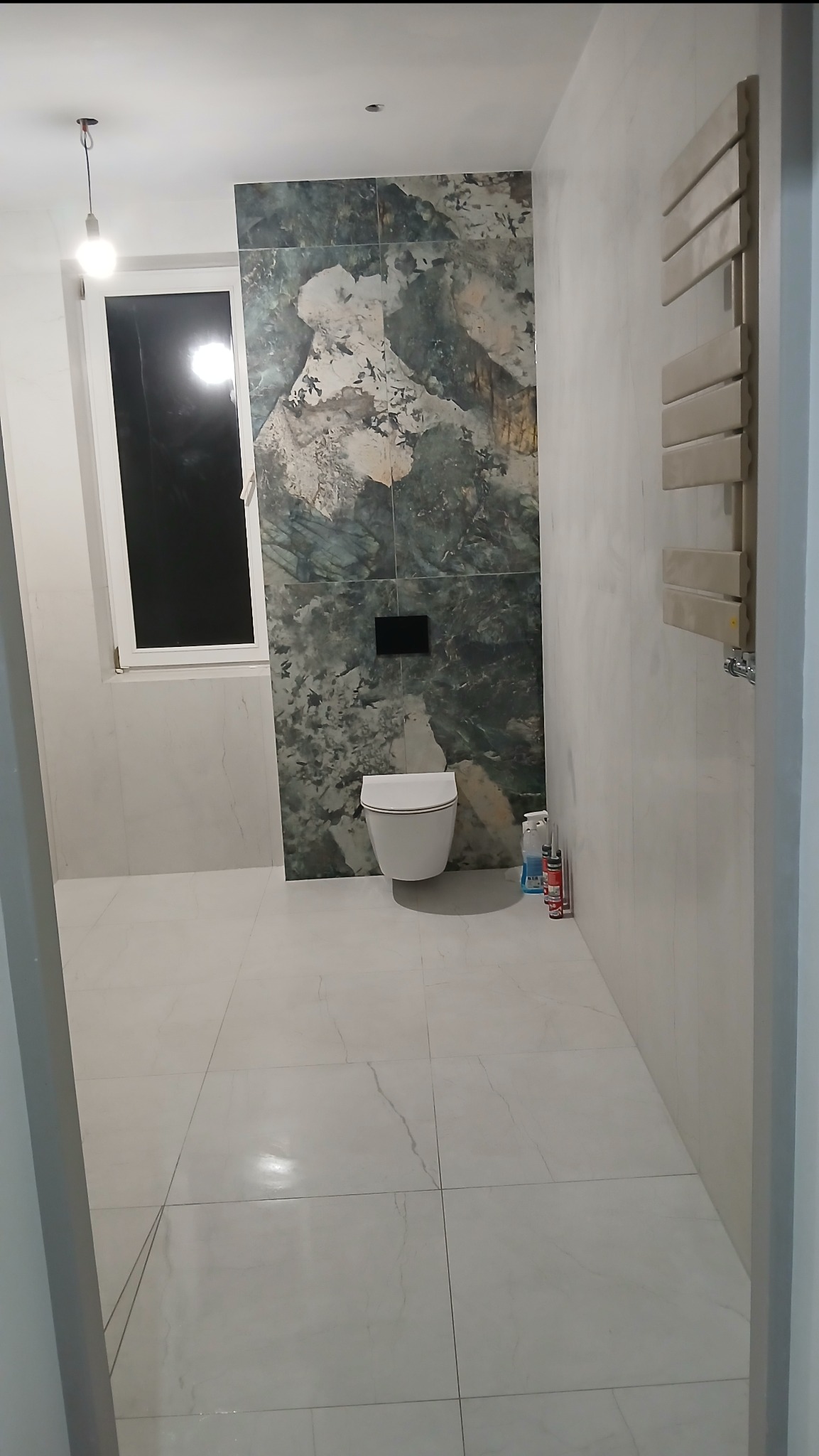 Wykończona łazienka z marmurową ścianą za toaletą, białe płytki podłogowe i grzejnik drabinkowy. Widoczne akcesoria montażowe i okno.