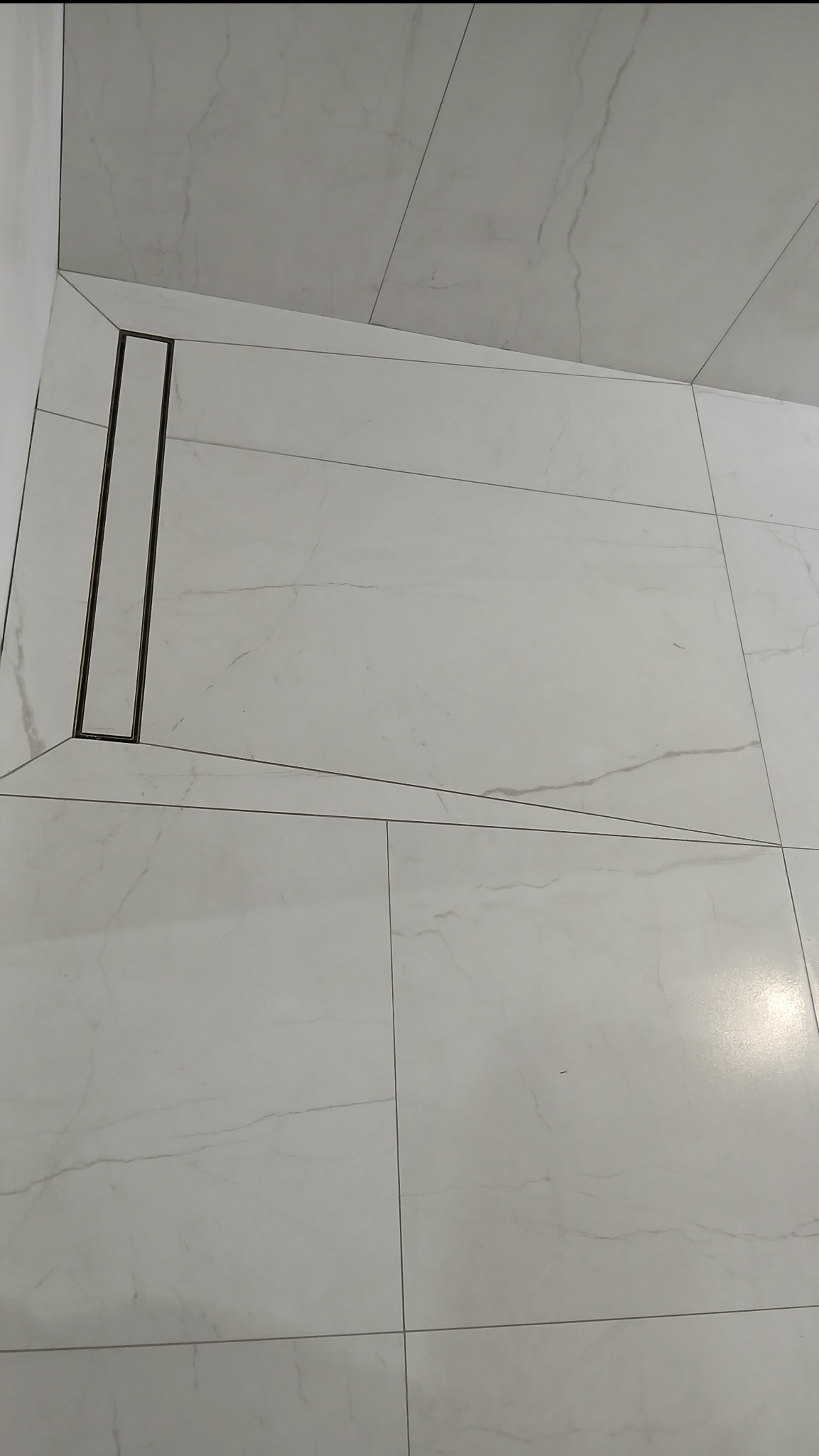 Białe, marmurowe płytki podłogowe w łazience z odpływem liniowym. Ułożenie płytek tworzy geometryczny wzór. Widok z góry, oświetlenie naturalne.