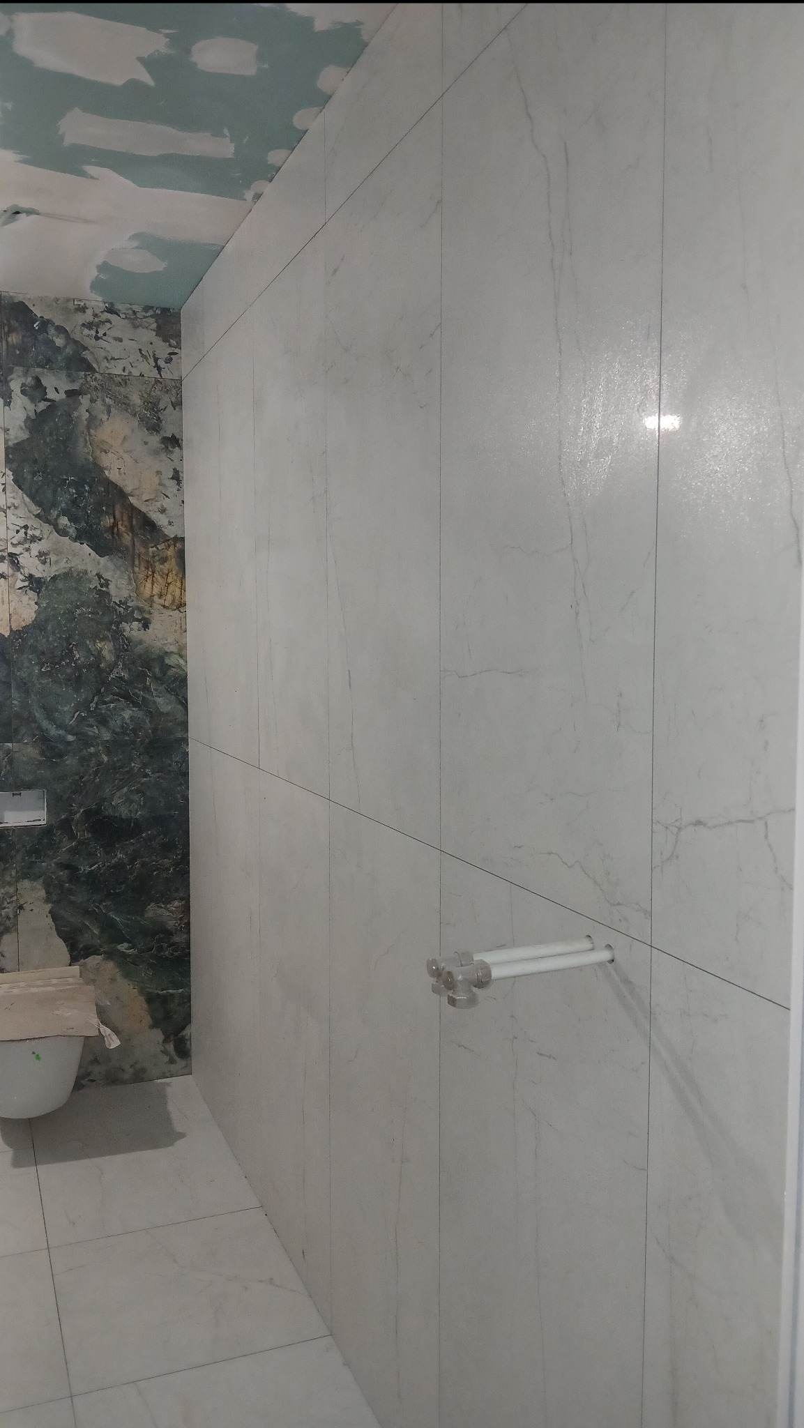 Wykończona łazienka z marmurowymi płytkami na ścianach i podłodze. Widoczny fragment toalety i uchwyt na ręczniki. Sufit w trakcie prac wykończeniowych.