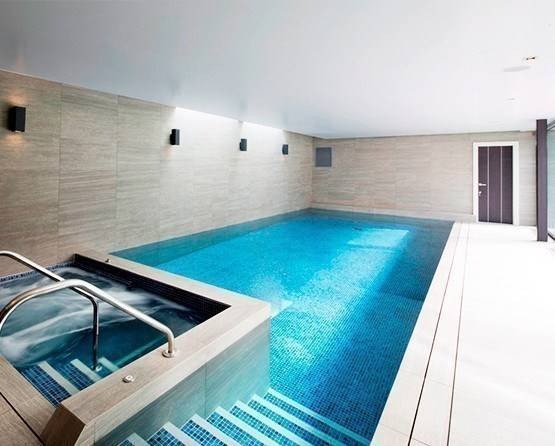 Nowoczesny basen kryty z jacuzzi, wykończony płytkami w odcieniach błękitu i szarości, oświetlony punktowymi lampami ściennymi i światłem naturalnym.