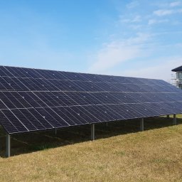 GM SYSTEM GRZEGORZ MIROSŁAW - Panel słoneczny na metalowej konstrukcji na trawiastym terenie, w tle dom w trakcie budowy i błękitne niebo.