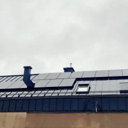 GM SYSTEM GRZEGORZ MIROSŁAW - Ciemna, metalowa dachówka z panelami słonecznymi, kominami i oknami dachowymi na tle pochmurnego nieba.