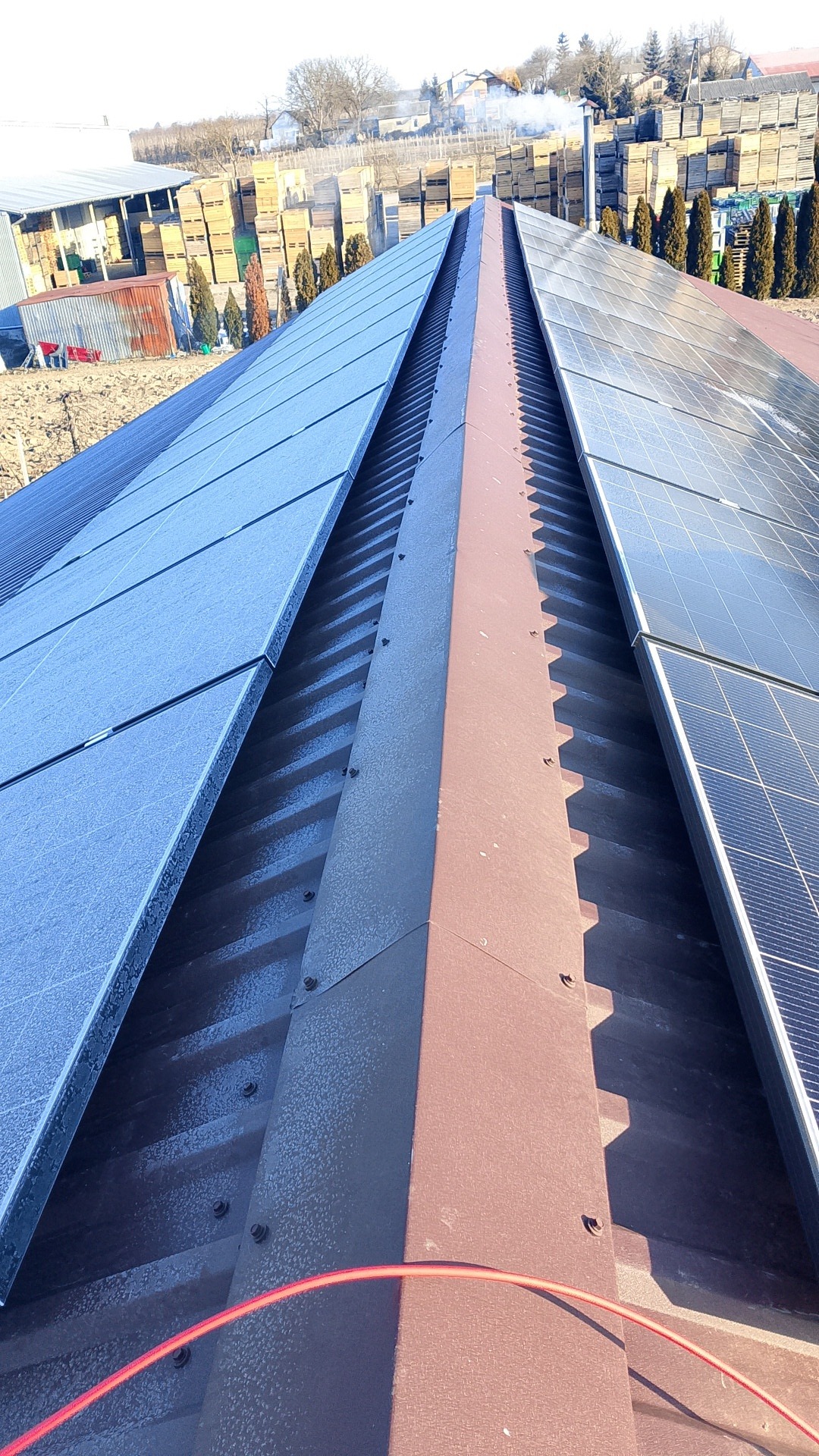 Widok z góry na panele słoneczne zamontowane na dachu z blachy trapezowej, z widoczną linią kalenicy i elementami mocującymi. W tle budynki gospodarcze i drzewa.