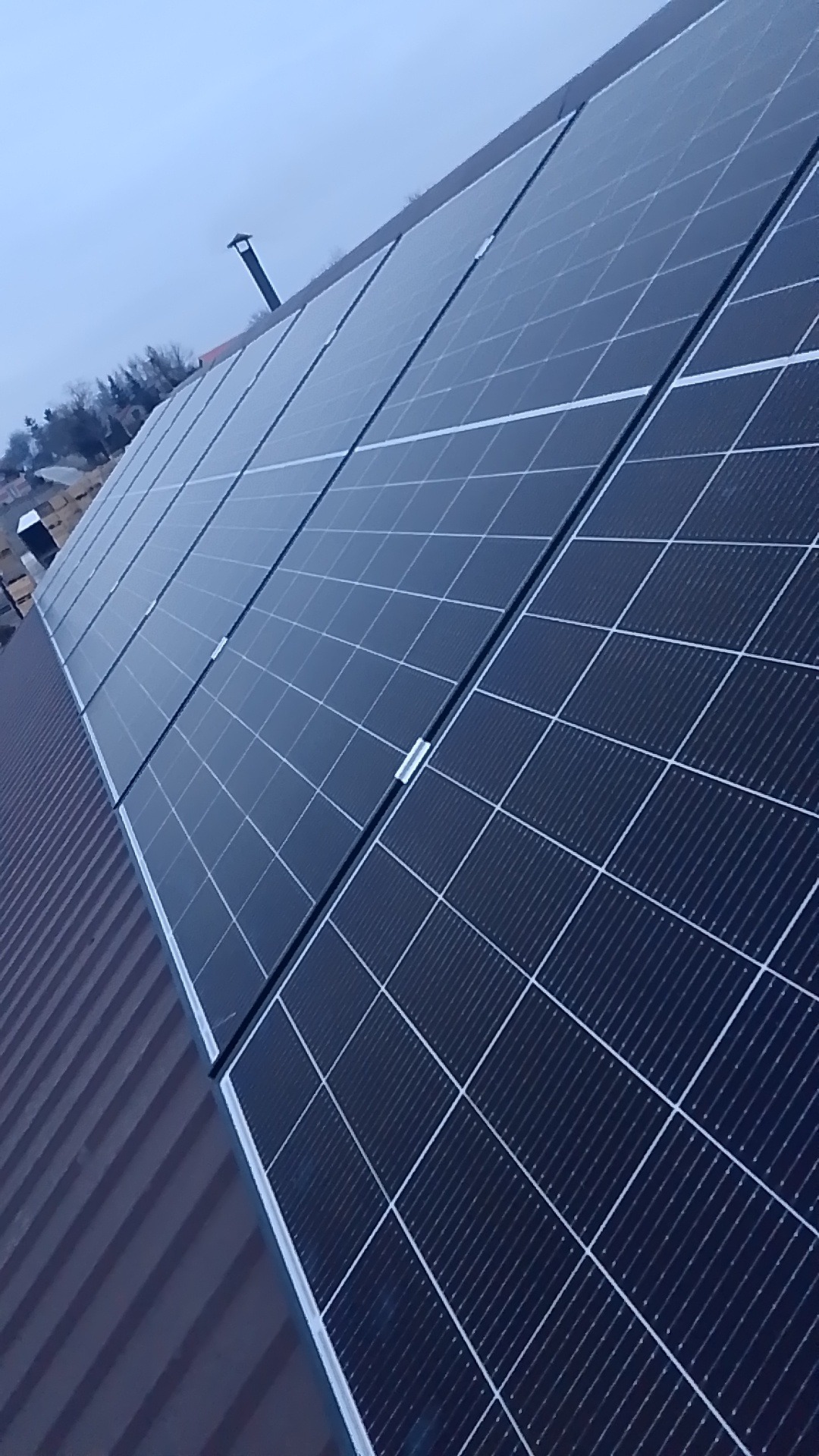 Rząd paneli fotowoltaicznych zamontowanych na metalowym dachu skośnym, widok z dołu pod kątem, w tle komin i zabudowania.