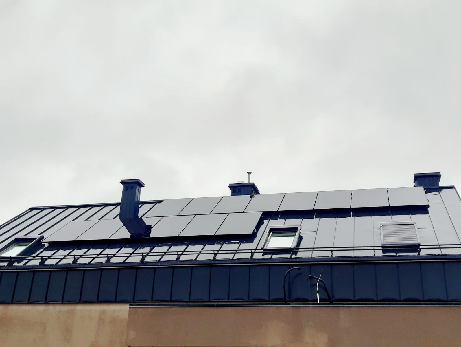 Ciemna, metalowa dachówka z panelami słonecznymi, kominami i oknami dachowymi na tle pochmurnego nieba.