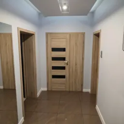 Korytarz z brązowymi płytkami podłogowymi, drewnianymi drzwiami z czarnymi, poziomymi paskami i oświetleniem sufitowym