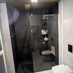 RAD-ROL - Nowoczesna łazienka z kabiną prysznicową typu walk-in, czarnymi płytkami pod prysznicem i szarymi płytkami na podłodze, drewnopodobnym sufitem oraz podwieszaną toaletą.
