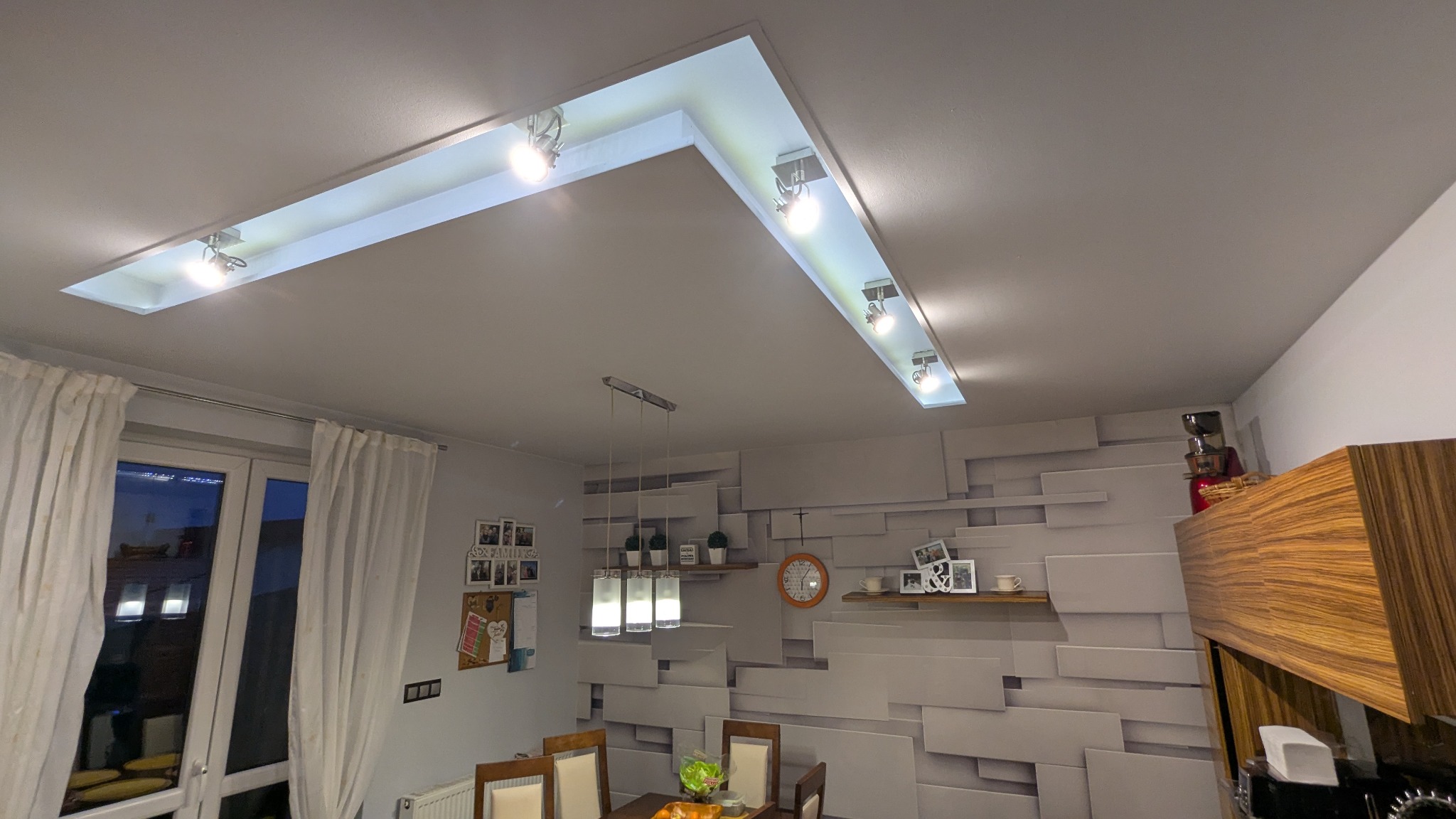 Nowoczesny sufit podwieszany z oświetleniem punktowym w kształcie litery L, widok na stół jadalny i dekoracyjną ścianę z geometrycznym wzorem.