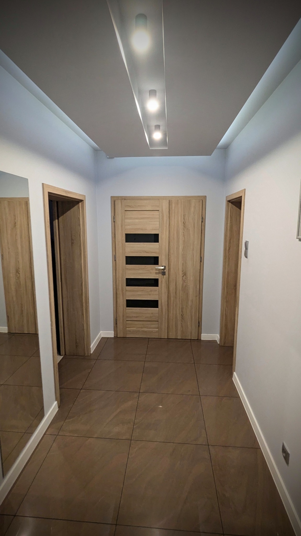 Korytarz z brązowymi płytkami podłogowymi, drewnianymi drzwiami z czarnymi, poziomymi paskami i oświetleniem sufitowym