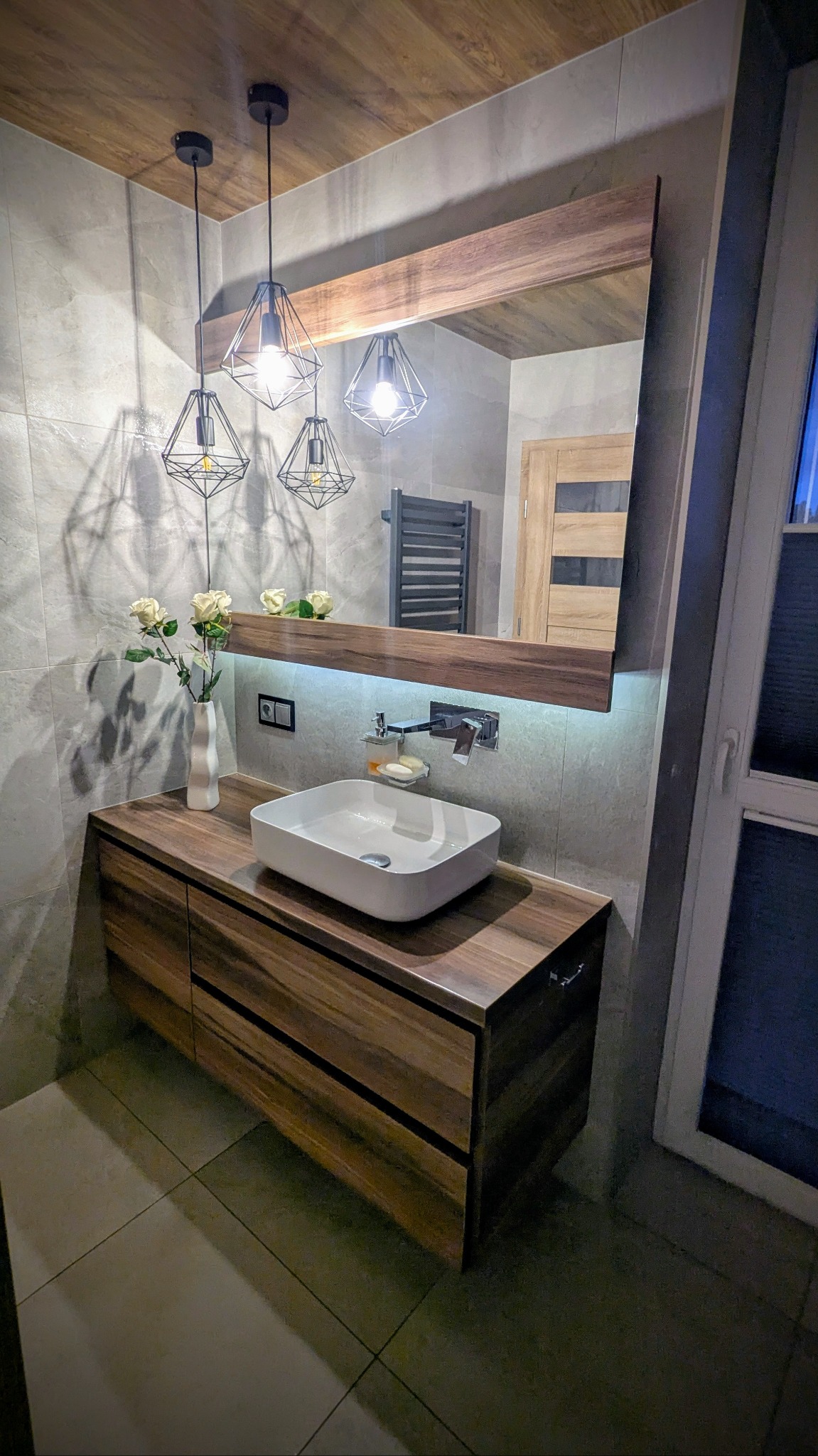 Nowoczesna łazienka z drewnianą szafką pod umywalką, lustrem z podświetleniem LED i geometrycznymi lampami wiszącymi.