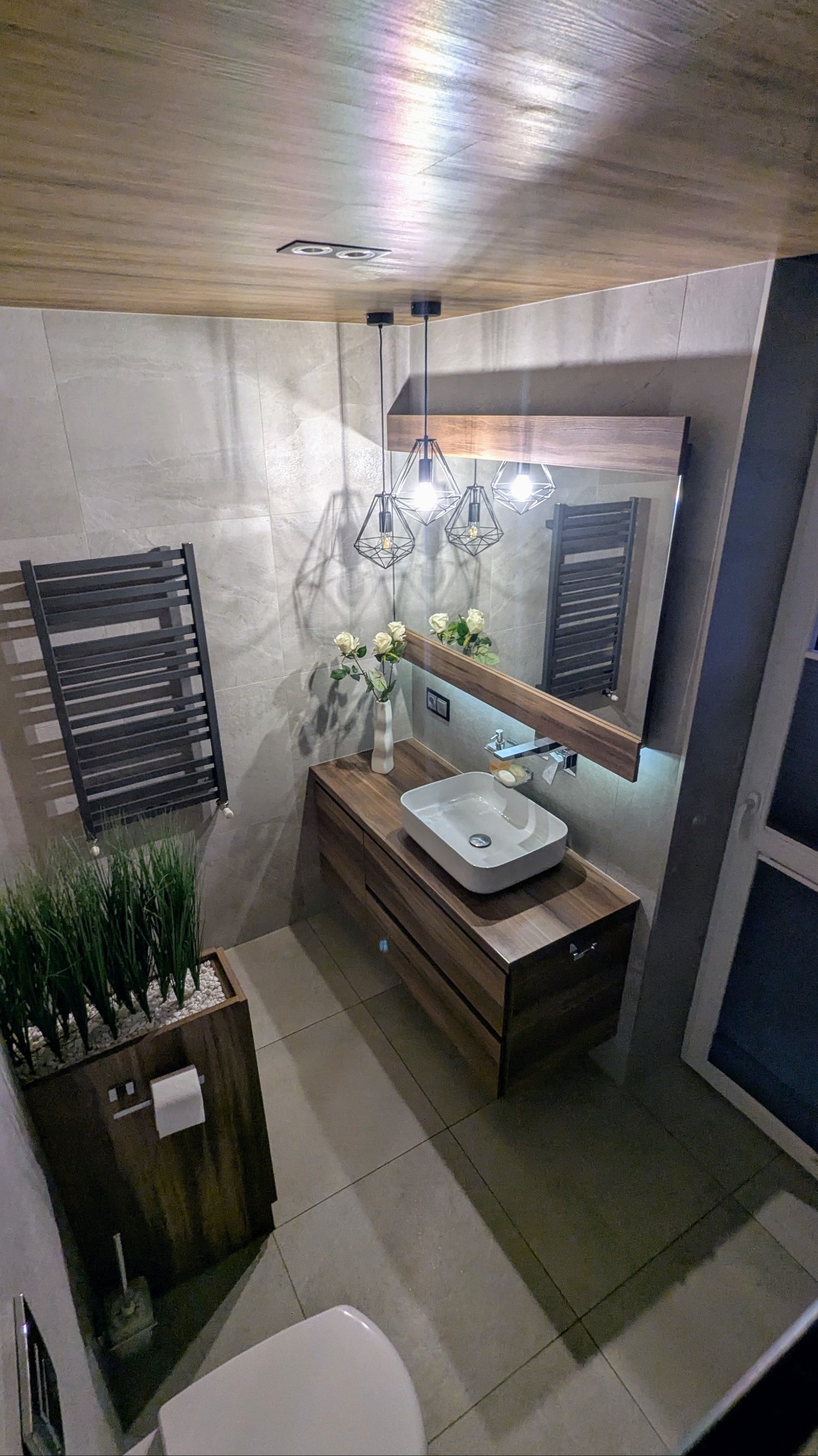 Nowoczesna łazienka z drewnianymi elementami: szafka pod umywalkę, rama lustra i sufit. Umywalka nablatowa z armaturą, dekoracyjne lampy wiszące, grzejnik drabinkowy i roślina w drewnianej donicy.
