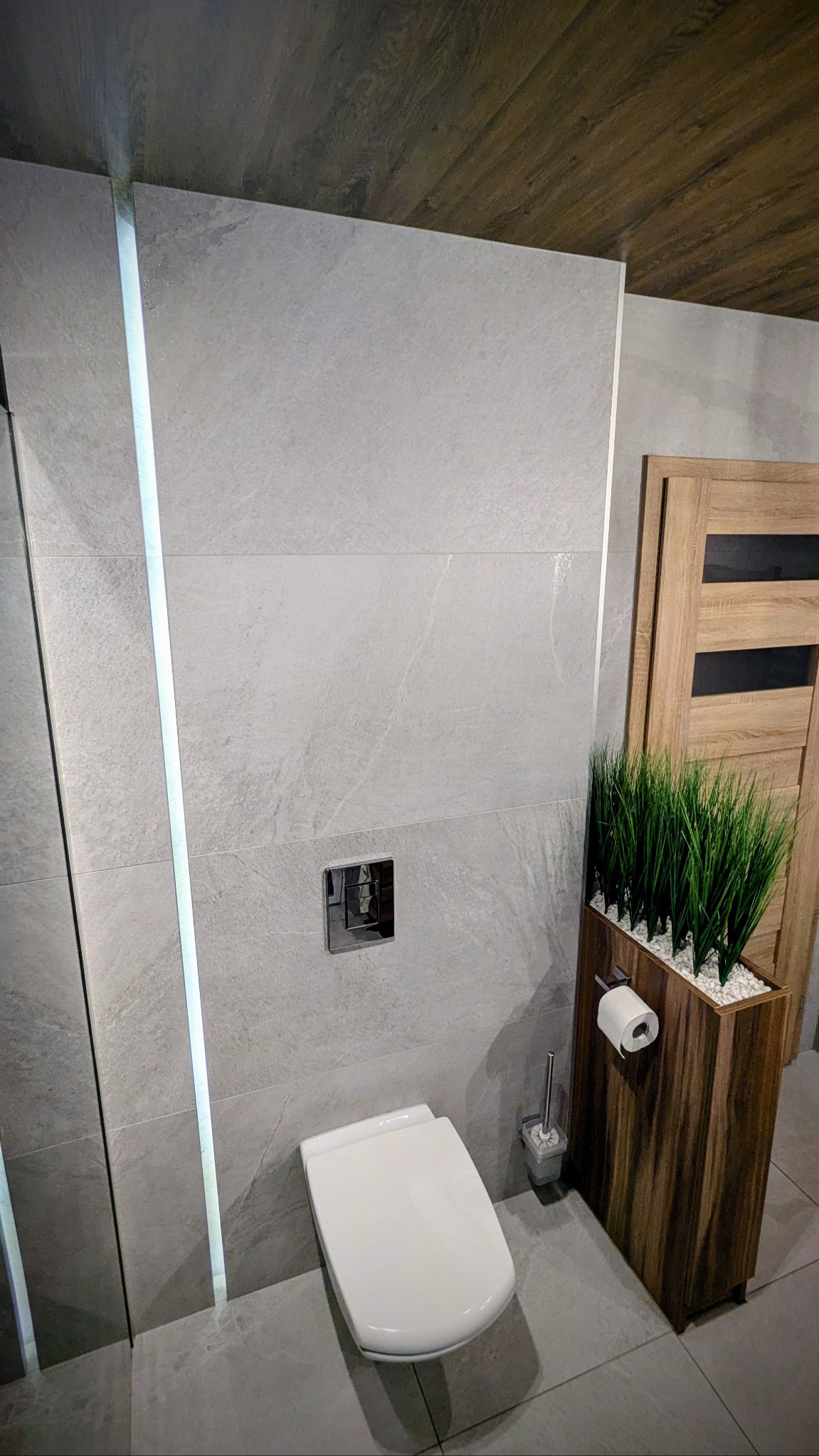 Nowoczesna łazienka z szarymi płytkami imitującymi kamień, podświetlanymi pionowymi listwami LED, wiszącą toaletą, chromowanym przyciskiem spłukującym oraz drewnianą donicą z ozdobną trawą...