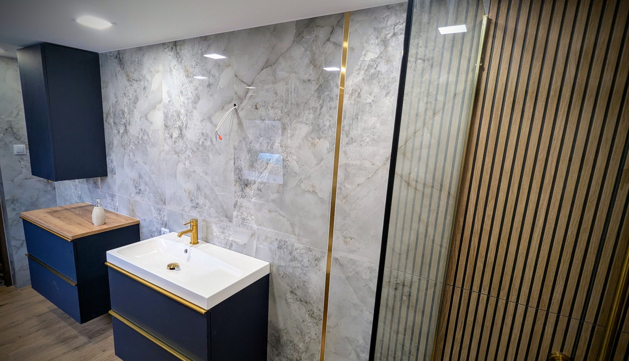 Nowoczesna łazienka z marmurowymi płytkami, złotymi akcentami, umywalką nablatową i drewnianą ścianką działową z listew.