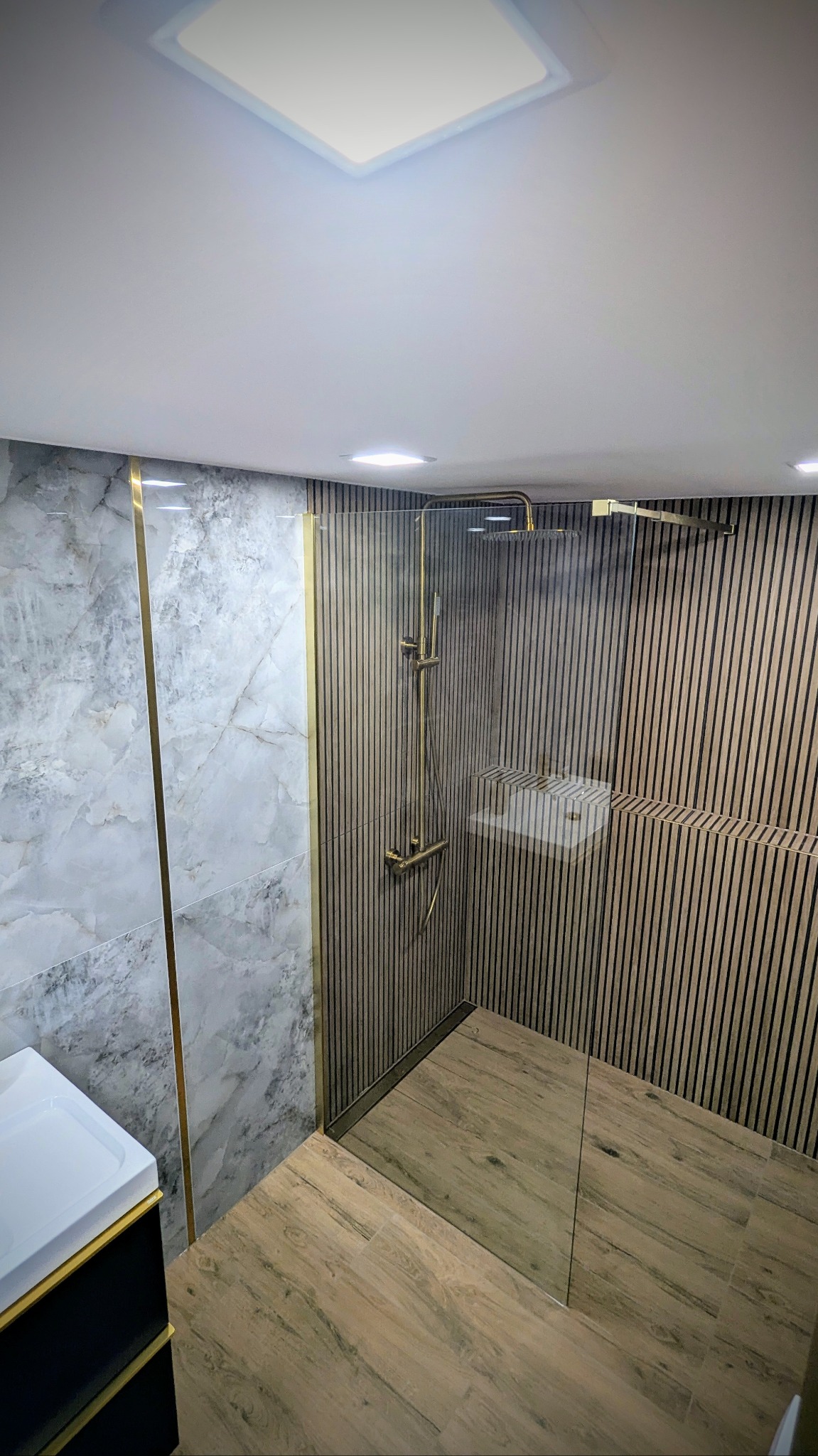 Nowoczesna łazienka z marmurową ścianą, złotymi akcentami i kabiną prysznicową typu walk-in z drewnianymi panelami ściennymi.