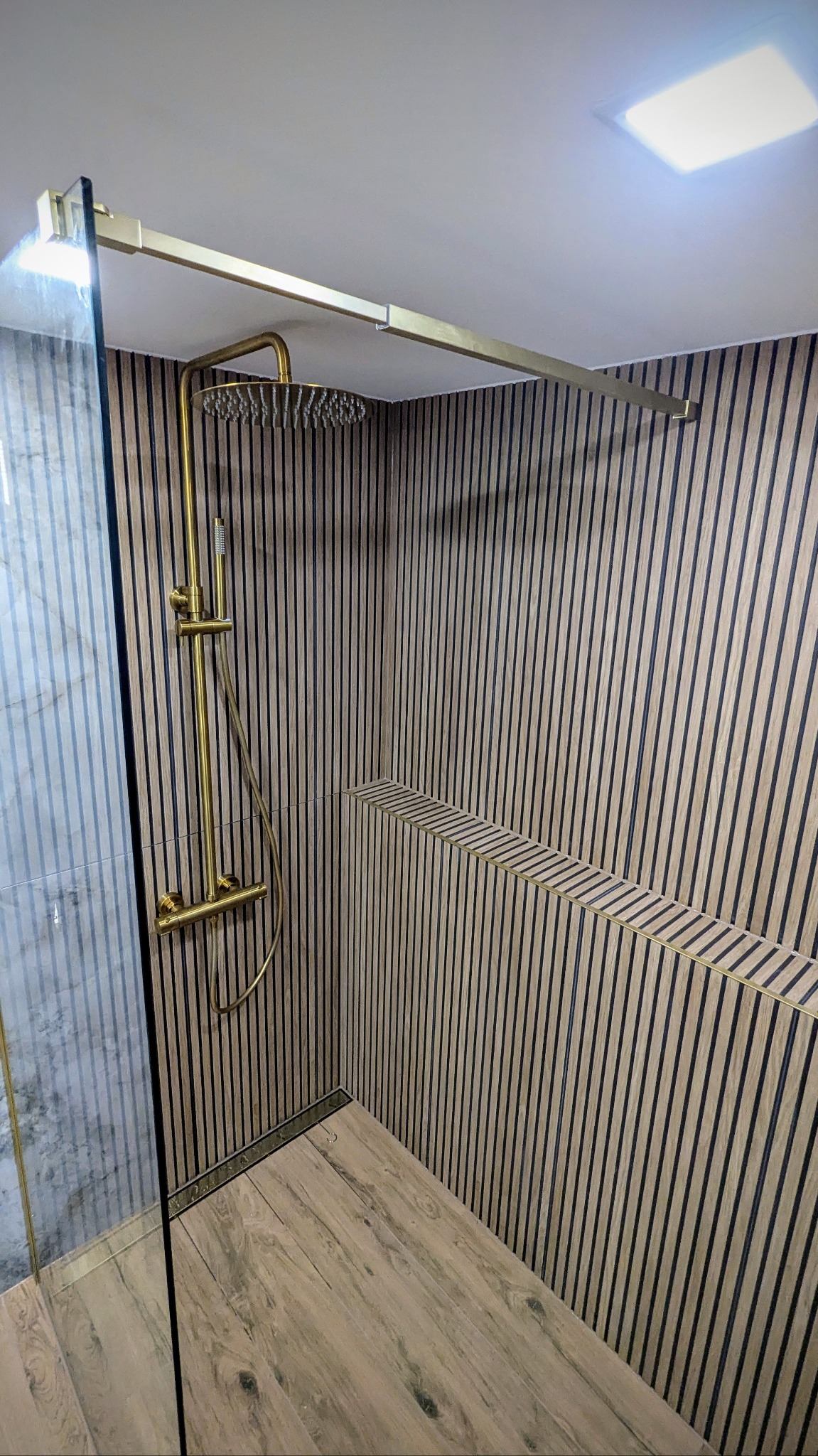 Nowoczesna kabina prysznicowa z deszczownicą w kolorze złotym, wykończona panelami ściennymi w pionowe paski i drewnopodobną podłogą, z odpływem liniowym.