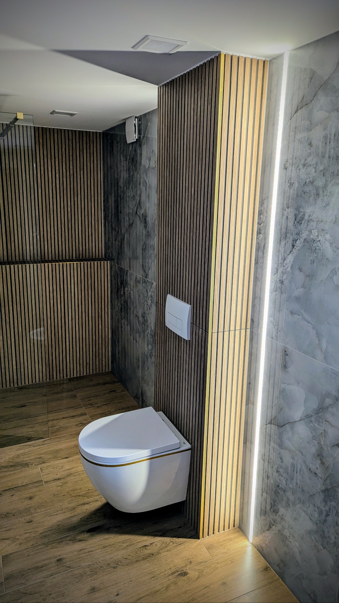 Nowoczesna łazienka z podwieszaną toaletą, drewnianymi lamelami na ścianie z oświetleniem LED i marmurowymi płytkami.