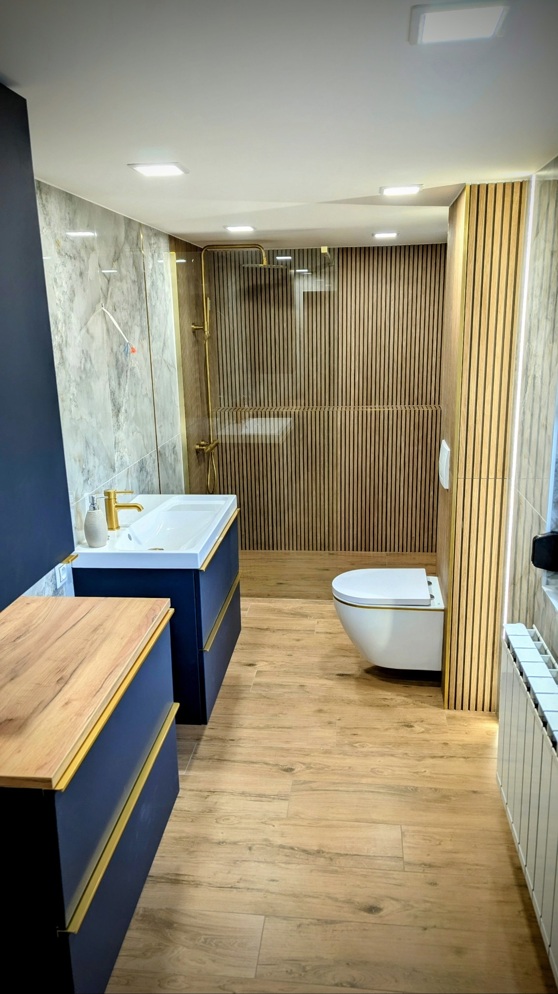 Nowoczesna łazienka z drewnianymi lamelami na ścianie prysznica, ciemnoniebieską szafką pod umywalką z złotymi akcentami i drewnopodobną podłogą z płytek.