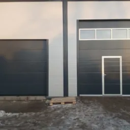 Dwie szare bramy segmentowe w hali przemysłowej, jedna z oknami i drzwiami, paleta drewniana u podstawy mniejszej bramy, resztki śniegu na ziemi.