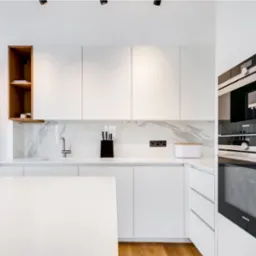 Nowoczesna, minimalistyczna kuchnia z białymi szafkami, marmurowym blatem i drewnianymi akcentami. Widoczny piekarnik Siemens i grzejnik dekoracyjny.