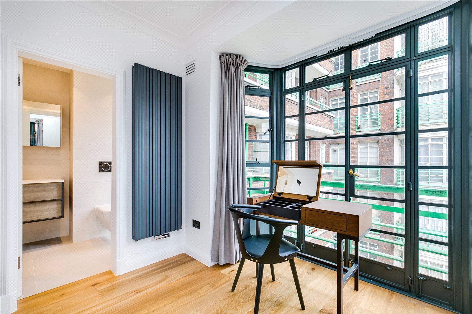 Jasne wnętrze z drewnianą podłogą, grzejnikiem panelowym, toaletką przy oknie i widokiem na łazienkę. Minimalistyczny design z nutą klasyki.