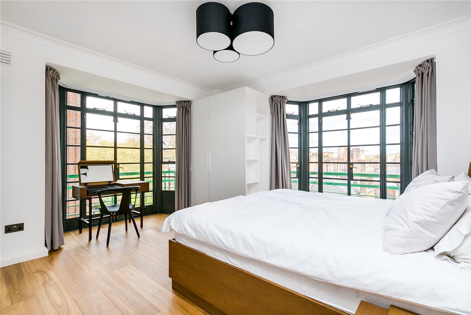 Jasna sypialnia z parkietem, dużym łóżkiem, biurkiem przy oknie i szafą wnękową. Nowoczesne oświetlenie sufitowe i szare zasłony dodają elegancji.