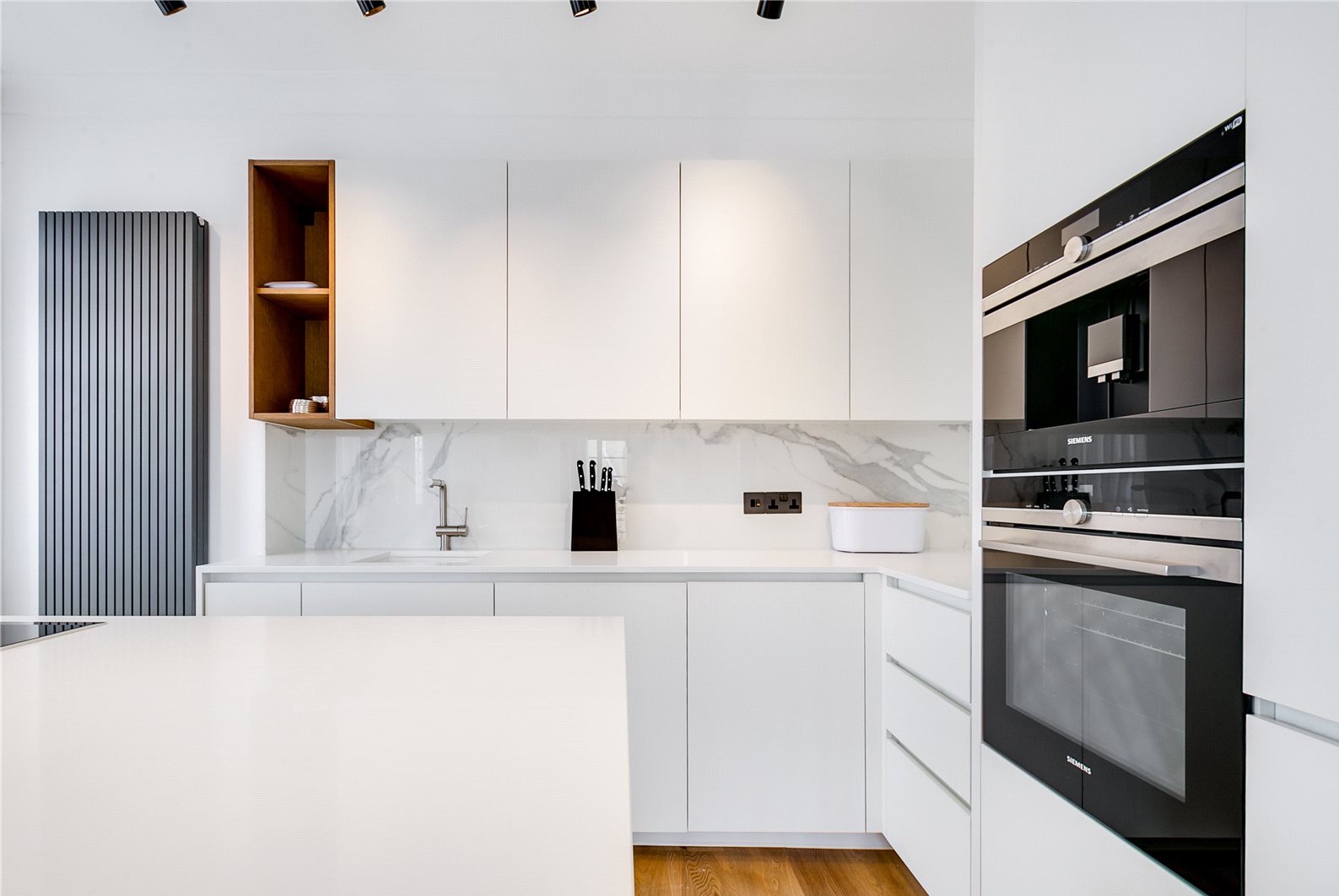 Nowoczesna, minimalistyczna kuchnia z białymi szafkami, marmurowym blatem i drewnianymi akcentami. Widoczny piekarnik Siemens i grzejnik dekoracyjny.
