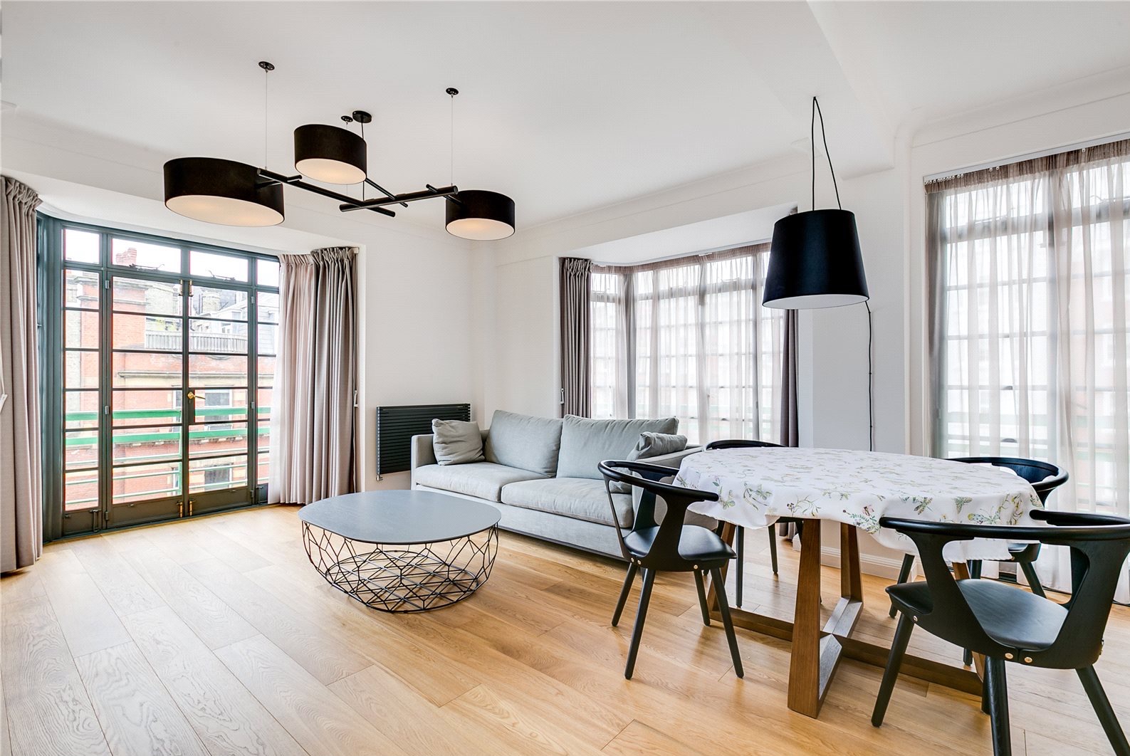 Jasne, nowoczesne wnętrze z drewnianą podłogą, szarą sofą i stolikiem kawowym z metalową podstawą. Okna z widokiem na miasto i designerskie lampy.