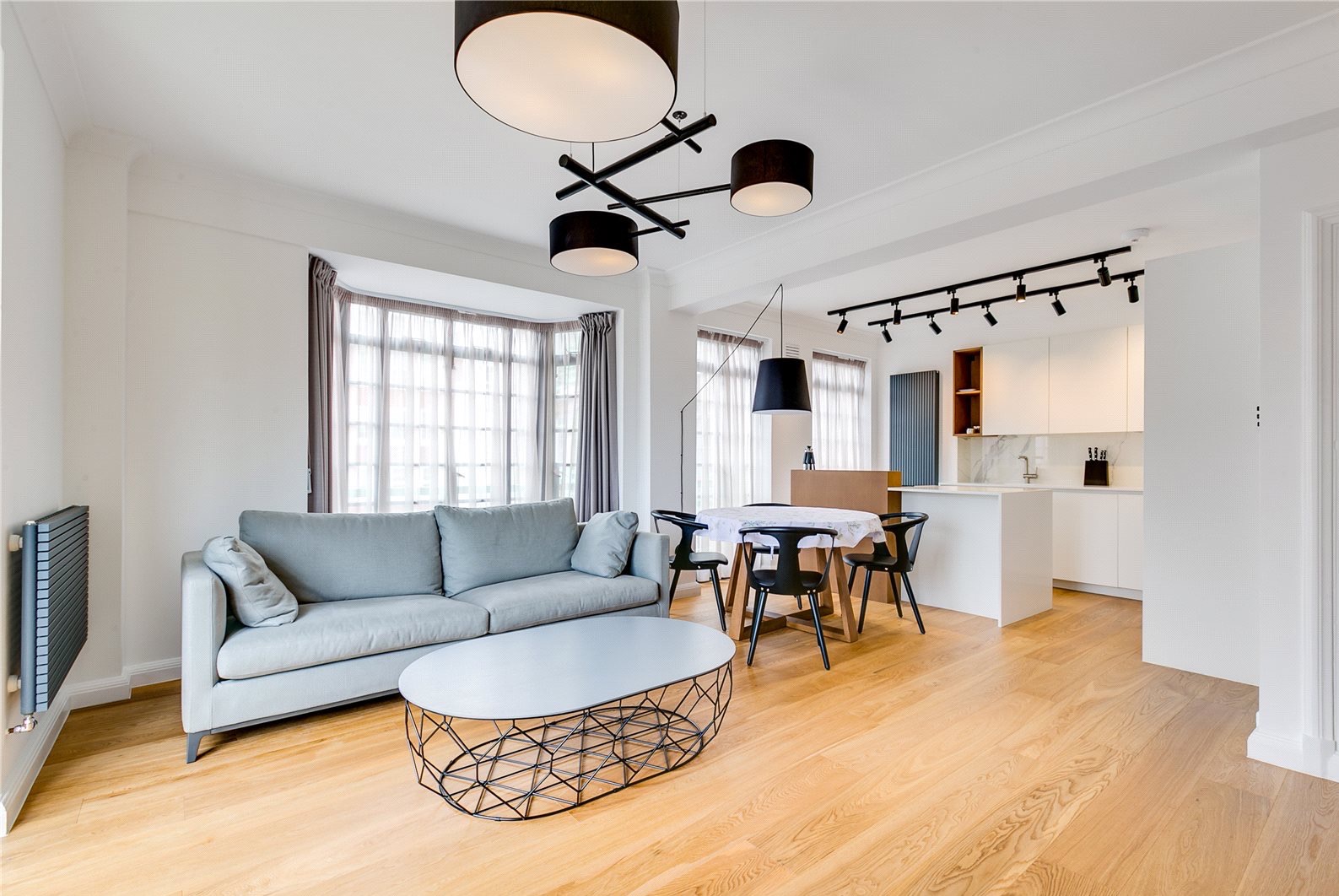 Jasne wnętrze po remoncie: salon z szarą sofą, stolikiem kawowym i jadalnią z kuchnią w tle. Nowoczesne oświetlenie sufitowe, drewniana podłoga i duże okno.