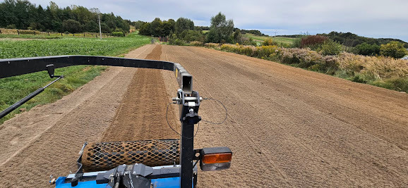 Widok z perspektywy operatora na maszynę do siewu trawy, pracującą na dużej powierzchni pola. W tle widoczne zielone pola uprawne i drzewa pod pochmurnym niebem.