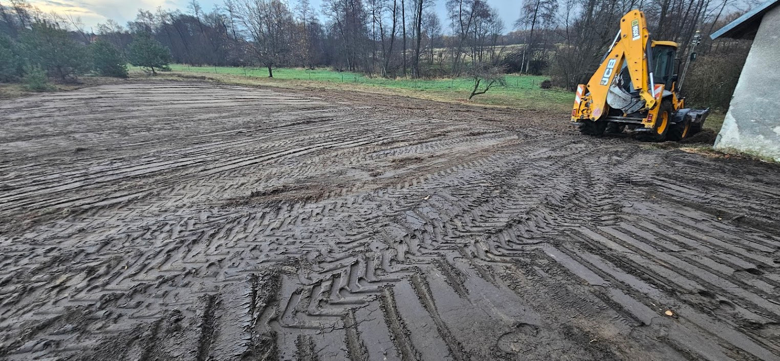 Żółta koparko-ładowarka JCB na błotnistym terenie z wyraźnymi śladami opon, przygotowanym pod przyszłe prace ogrodowe, w tle drzewa i budynek.