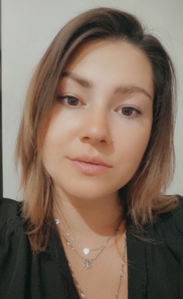 Portret młodej kobiety z brązowymi włosami, noszącej czarną bluzkę i srebrny naszyjnik z zawieszkami w kształcie serca i czterolistnej koniczyny. Ma piercing w nosie.