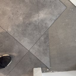 EUROBUD-SK Serhii Kasianchuk - Układanie szarych, kwadratowych płytek podłogowych, widoczny fragment buta, miarka, ołówek i biała listwa przypodłogowa.