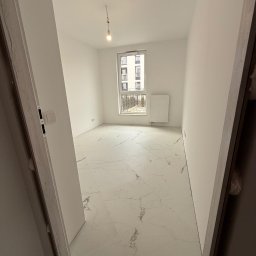 EUROBUD-SK Serhii Kasianchuk - Pusty, jasny pokój z białymi ścianami, marmurową podłogą i grzejnikiem pod oknem, widok zza otwartych drzwi z surową ościeżnicą.