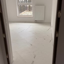 EUROBUD-SK Serhii Kasianchuk - Jasne płytki podłogowe imitujące marmur w nowym, pustym pomieszczeniu z oknem i grzejnikiem, widok przez framugę drzwi z widoczną pianką montażową.