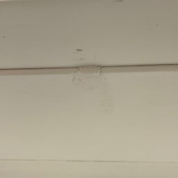 EUROBUD-SK Serhii Kasianchuk - Fragment białej, pomalowanej powierzchni z odpryskiem farby. Widoczna nierówna faktura i ślady po naprawie. Detal uszkodzenia powłoki malarskiej.