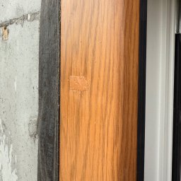 EUROBUD-SK Serhii Kasianchuk - Fragment okna z drewnopodobną okleiną, na której widać ślady naprawy. Obok surowy beton i czarna taśma uszczelniająca. Detal okna po renowacji.