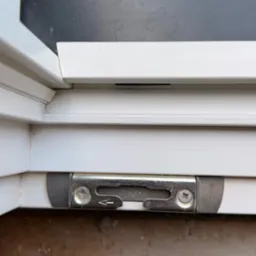 Szczegółowe ujęcie mechanizmu blokującego w białej ramie okiennej z widocznymi śrubami i strzałką kierunkową na metalowym elemencie, ukazujące precyzję montażu.