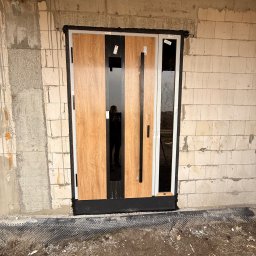 EUROBUD-SK Serhii Kasianchuk - Nowoczesne drzwi wejściowe z drewnianym panelem i czarnymi szybami, zamontowane w surowym murze z betonu komórkowego. Widoczne detale montażu i folia fundamentowa.