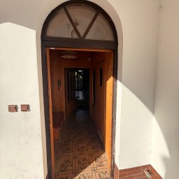 EUROBUD-SK Serhii Kasianchuk - Otwarty drewniany łuk drzwiowy z półokrągłym oknem w białej ścianie, widok na wnętrze z boazerią i wzorzystą podłogą. Cień słońca na ścianie.