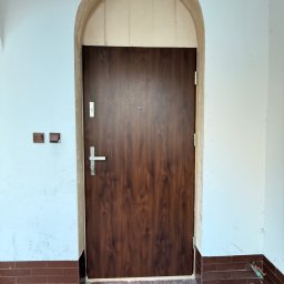 EUROBUD-SK Serhii Kasianchuk - Ciemne, drewnopodobne drzwi wejściowe z ościeżnicą w kształcie łuku, zamontowane w jasnym pomieszczeniu z brązowymi płytkami podłogowymi i ceglaną lamperią.