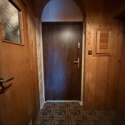 EUROBUD-SK Serhii Kasianchuk - Ciemne drzwi wejściowe z metalową klamką, osadzone w łukowatym przejściu, otoczone boazerią i wzorzystą podłogą. Widoczny fragment okna z fakturą szkła.