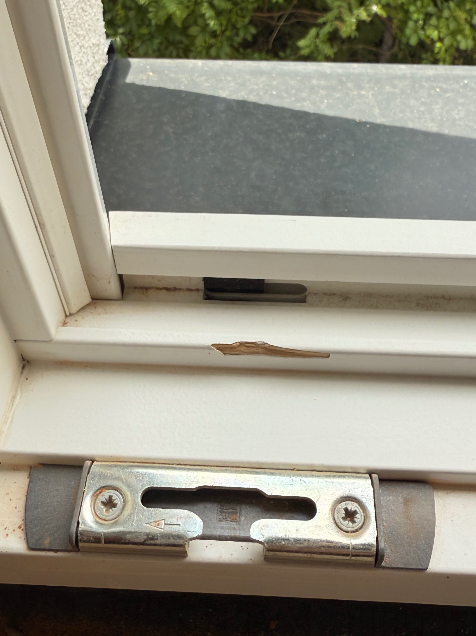 Uszkodzony biały profil okienny z widocznym pęknięciem i rdzawym nalotem. Metalowy element okucia z widocznymi śrubami i strzałką. Widok z bliska.