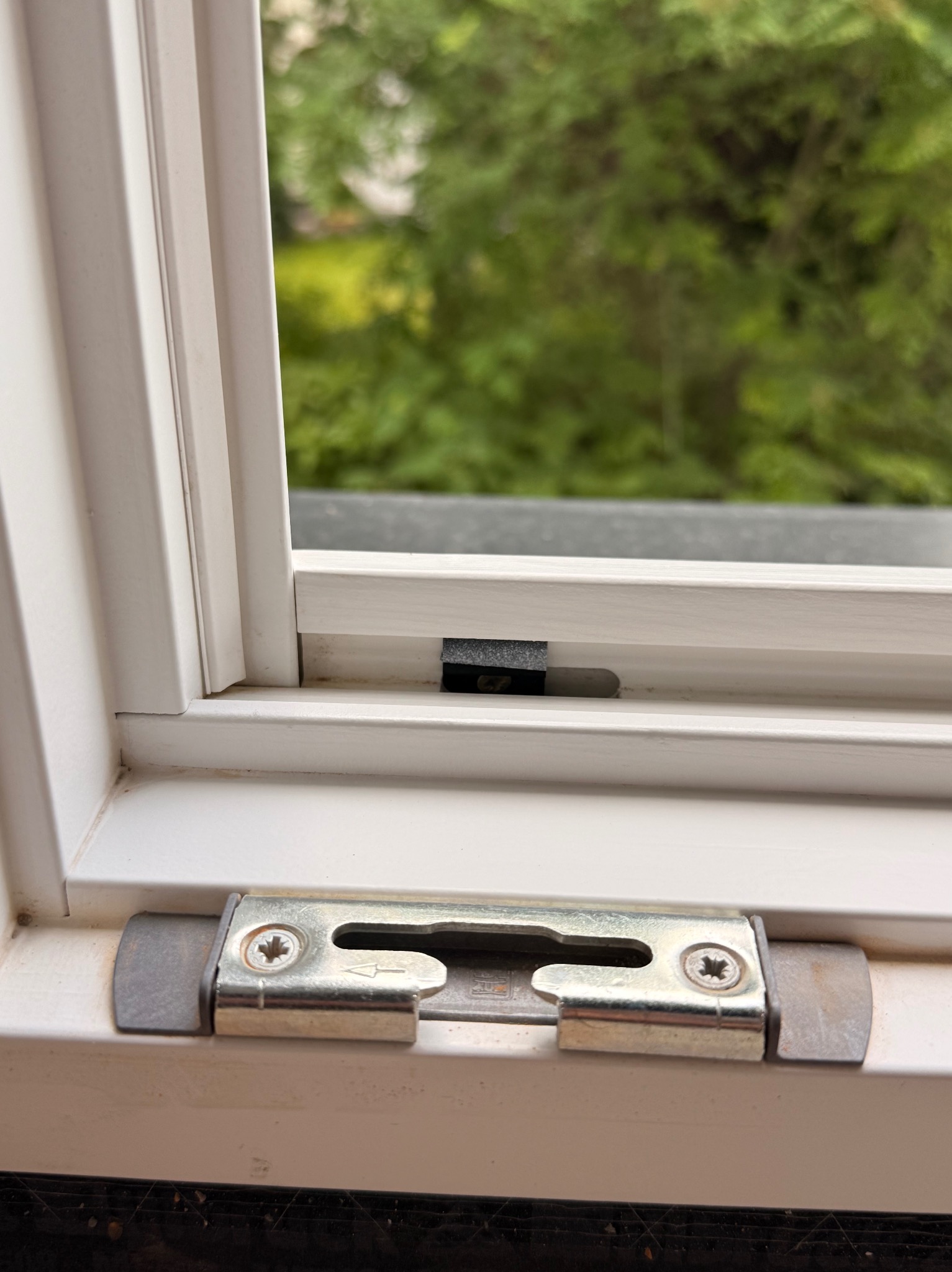 Białe okno PCV z widocznym mechanizmem blokującym, metalowym, z dwoma śrubami. W tle zieleń. Widoczne zabrudzenia na ramie okiennej.