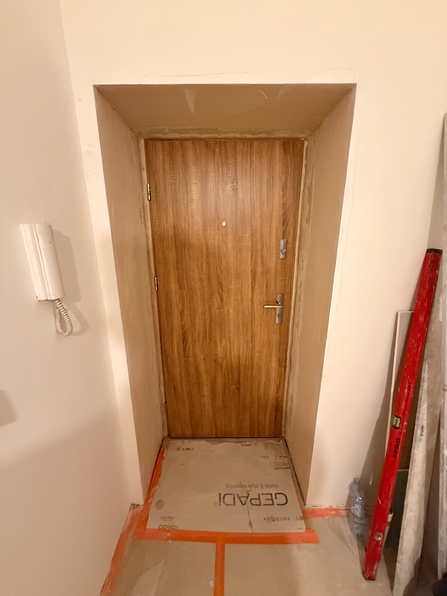 Montaż drzwi wewnętrznych z drewnopodobnym wykończeniem, widoczne ślady szpachlowania ościeżnicy, poziomica i zabezpieczenia podłogi.