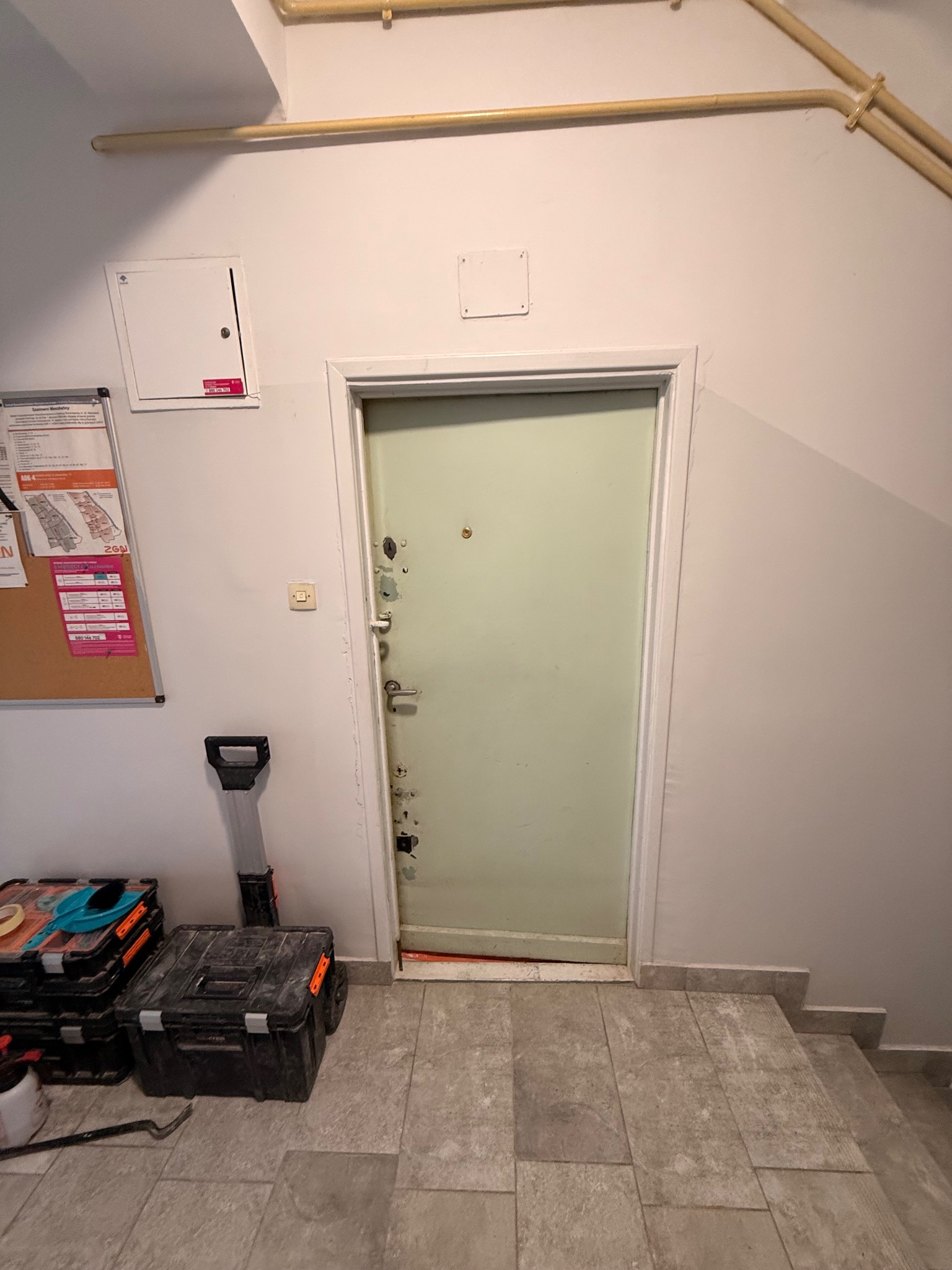 Drzwi wejściowe w bloku, widoczne ślady użytkowania, obok skrzynki z narzędziami. Ujęcie z dołu, widoczne rury pod sufitem i schody.