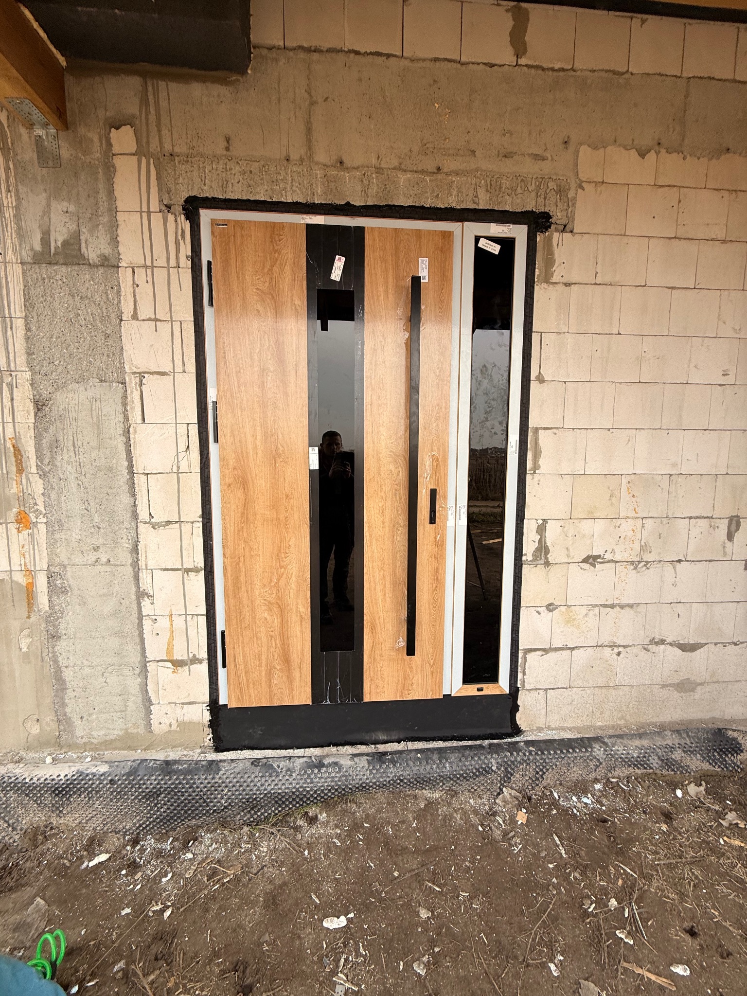 Nowoczesne drzwi wejściowe z drewnianym panelem i czarnymi szybami, zamontowane w surowym murze z betonu komórkowego. Widoczne detale montażu i folia fundamentowa.