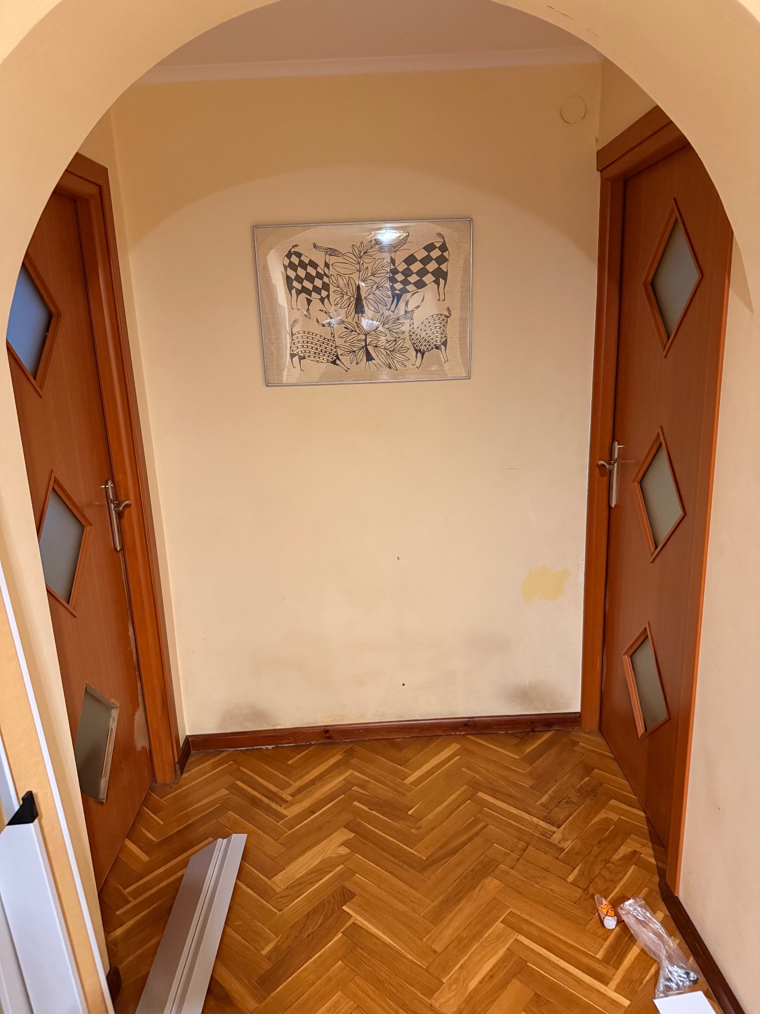 Korytarz z parkietem w jodełkę, dwoje drewnianych drzwi z szybkami w romby, obrazek na ścianie i listwy przypodłogowe do montażu. Widok na łukowate przejście.