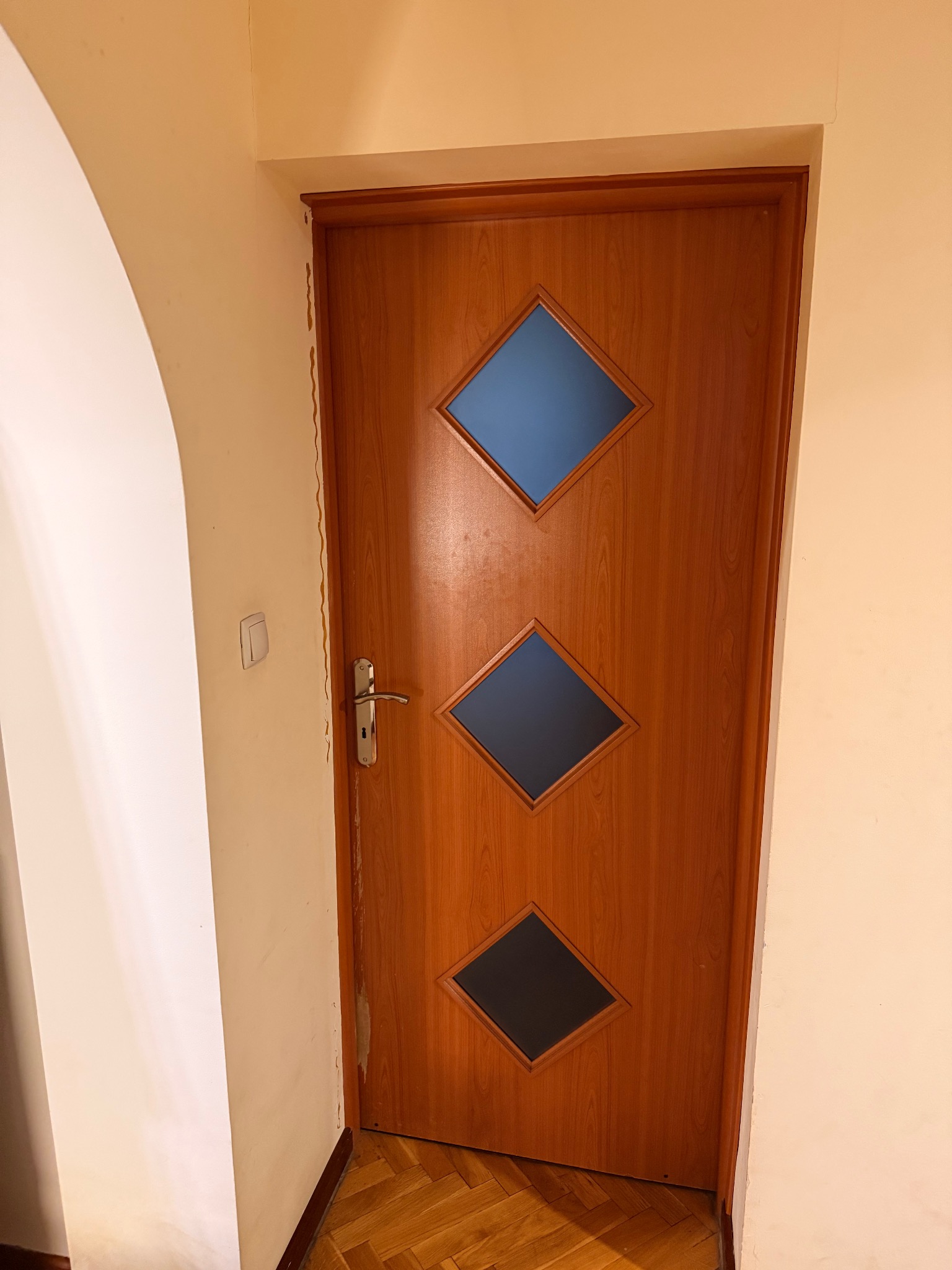 Drewniane drzwi wewnętrzne z trzema romboidalnymi przeszkleniami, widoczne ślady po malowaniu na ścianie obok ościeżnicy, klamka po lewej stronie.