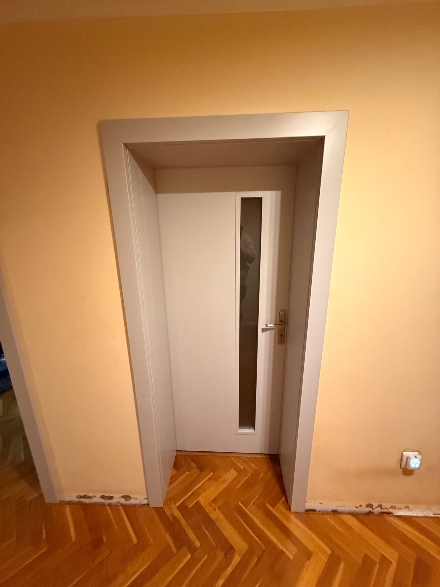 Białe drzwi z szybką w szarej ościeżnicy, zamontowane w ścianie o ciepłym, żółtym odcieniu. Parkiet ułożony w jodełkę. Widoczne ślady po remoncie przy podłodze.